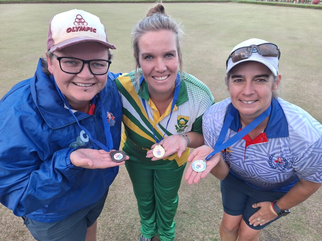Three women lawn bowlers standing together on a green, each holding up their medals and smiling.